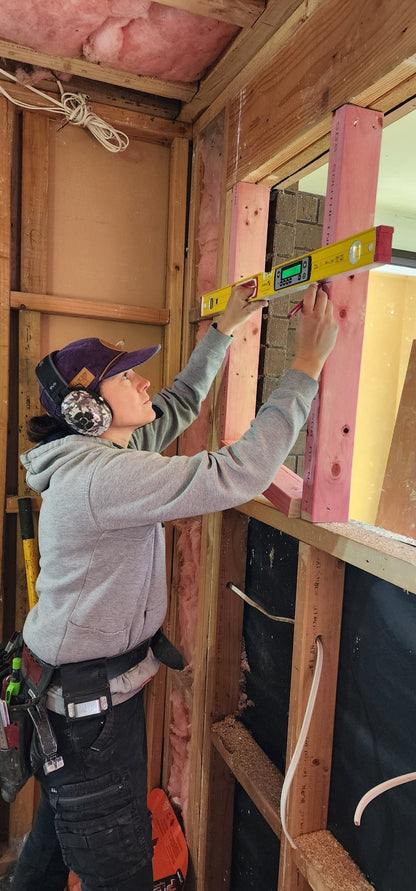 Female tradie checking wall framing with a level, wearing Mufftech Sole Bluetooth earmuffs